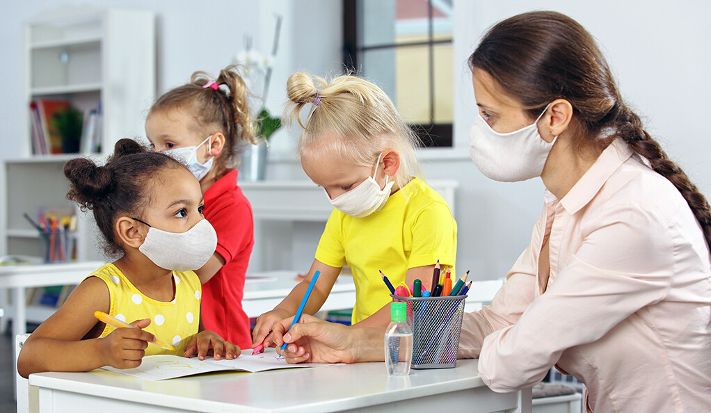 A teacher and kid with masks on their faces look into each other's eyes in class